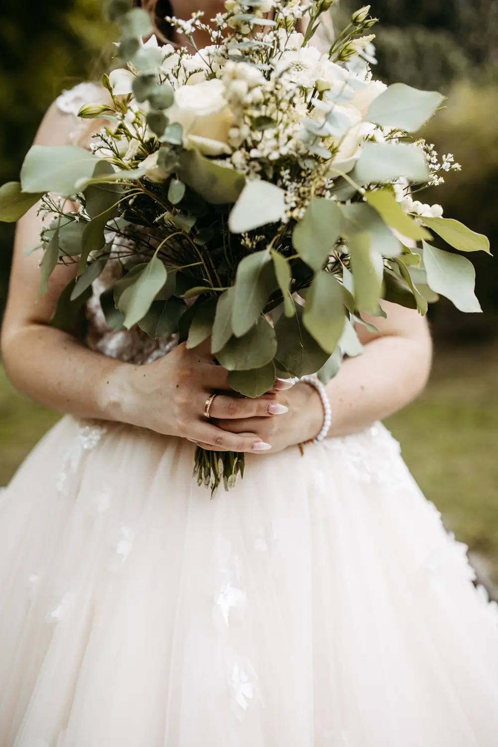 Eine Braut in einem weißen Hochzeitskleid hält einen großen Strauß mit weißen Blumen und Grünzeug. Nur ihr Oberkörper und ihre Hände sind sichtbar, mit einem Ring und einem Perlenarmband an ihrer linken Hand. Der Hintergrund besteht aus unscharfem Grün.