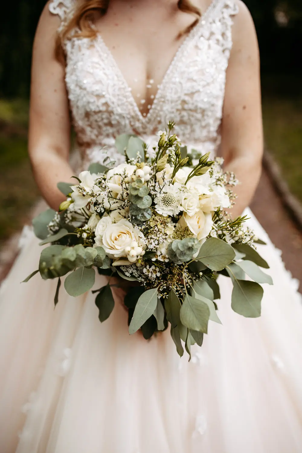 Eine Braut in einem eleganten Spitzenhochzeitskleid hält einen Strauß aus weißen Rosen, Grünzeug und kleinen Blumen und steht im Freien auf einem Weg. Nur ihr Oberkörper und ihr Strauß sind zu sehen.