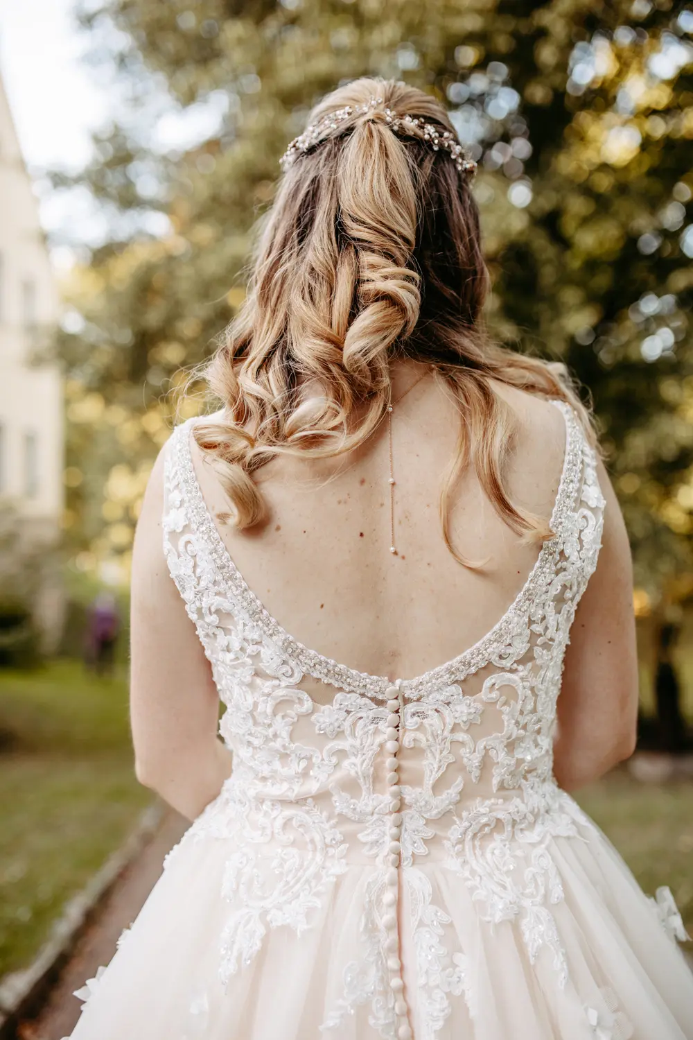 Eine Frau in einem weiĂźen, spitzenbestickten Hochzeitskleid wird von hinten gezeigt, mit langem, gewelltem Haar, das von einem zarten Haarschmuck geschmĂĽckt wird, im Freien inmitten von GrĂĽn.