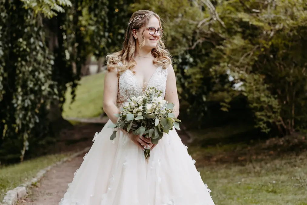 Eine Braut mit gewelltem Haar und Brille steht im Freien, lächelt und hält einen Strauß mit grünen und weißen Blumen. Sie trägt ein weißes, ärmelloses Hochzeitskleid mit vollem Rock und Spitzenverzierungen. Sie ist von Bäumen und Grünpflanzen umgeben.