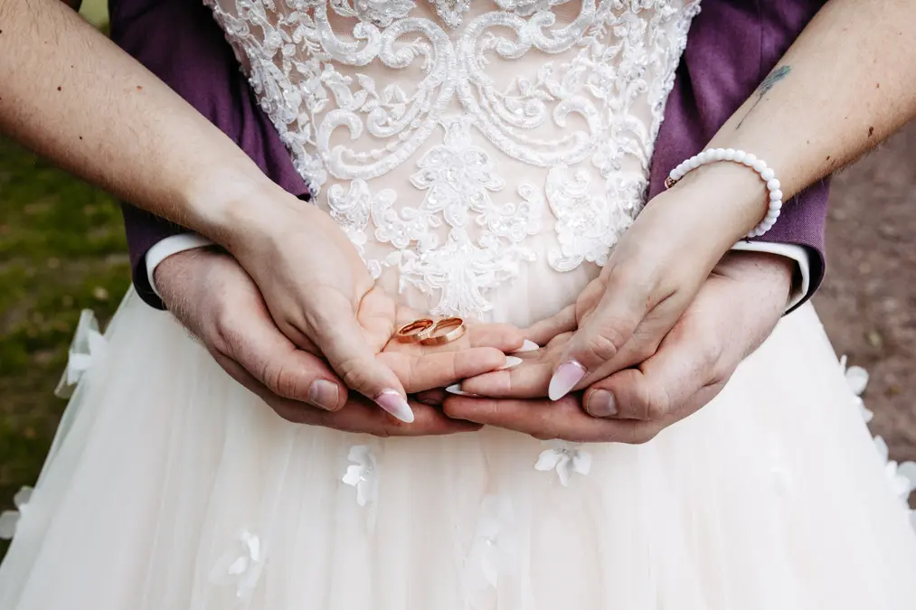 Nahaufnahme von Braut und Bräutigam, die zwei Eheringe in ihren Händen halten. Die Braut trägt ein weißes Spitzenkleid und ein Perlenarmband; die Hände des Bräutigams sind sanft um ihre gelegt.