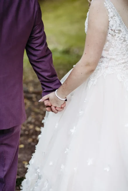 Ein Paar, das sich im Freien an den Händen hält; eine Person trägt einen lila Anzug, die andere ein weißes Hochzeitskleid aus Spitze mit Blumendetails. Nur ihre Torsos und Hände sind zu sehen.