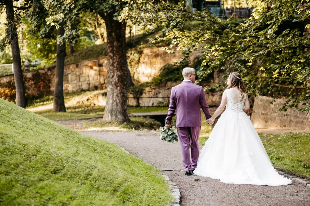 Eine Braut in einem weißen Kleid und ein Bräutigam in einem lilafarbenen Anzug gehen Hand in Hand einen von Bäumen gesäumten Weg in einem Park hinunter, umgeben von Grün und Sonnenlicht.