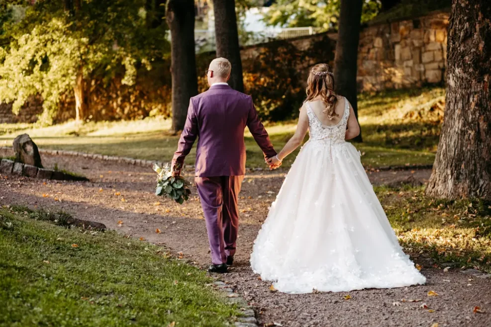 Eine Braut in einem weißen Kleid und ein Bräutigam in einem violetten Anzug gehen Hand in Hand einen von Bäumen gesäumten Parkweg entlang, während das Sonnenlicht durch die Bäume fällt.