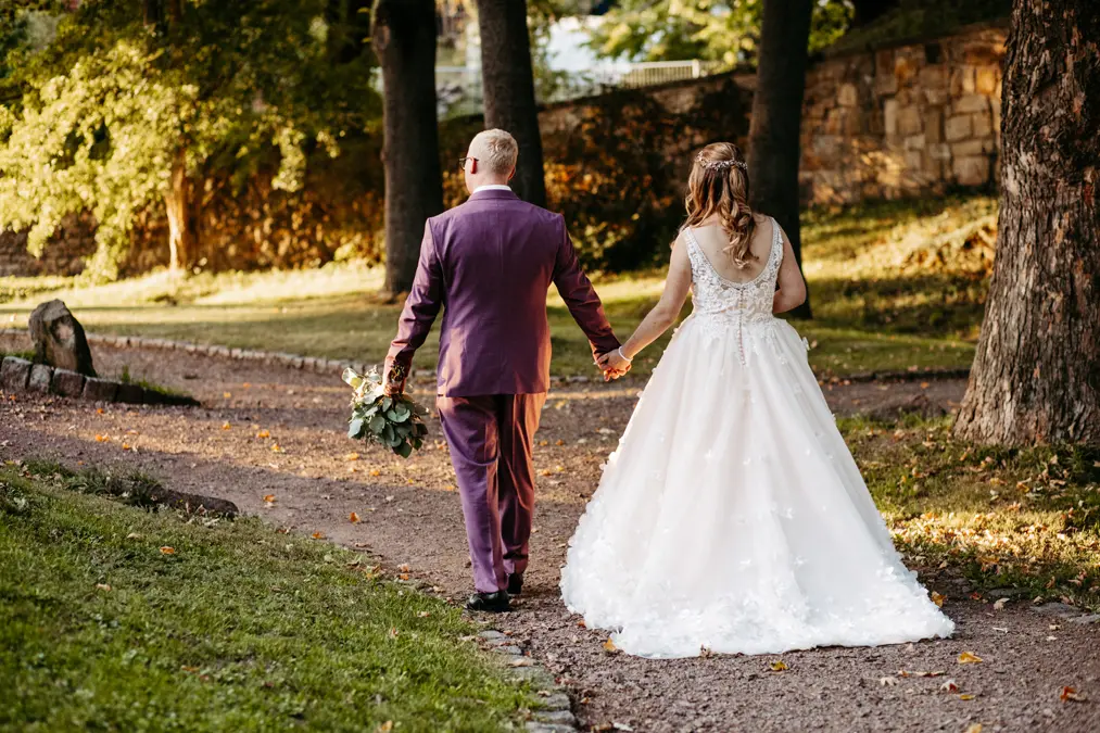 Eine Braut in einem weißen Kleid und ein Bräutigam in einem violetten Anzug gehen Hand in Hand einen von Bäumen gesäumten Parkweg entlang, während das Sonnenlicht durch die Bäume fällt.