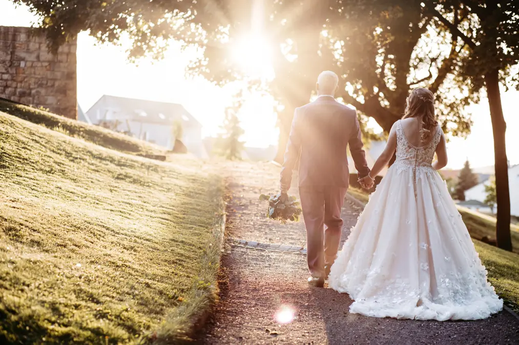 Eine Braut und ein Bräutigam gehen Hand in Hand einen sonnenbeschienenen Weg entlang, umgeben von grünem Gras und Bäumen. Die Braut trägt ein langes weißes Kleid, der Bräutigam einen Anzug. Das Sonnenlicht fällt durch die Bäume und schafft eine warme, romantische Atmosphäre.
