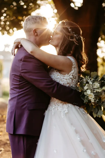 Eine Braut und ein Bräutigam küssen sich bei Sonnenuntergang im Freien, wobei das Sonnenlicht durch die Bäume hinter ihnen scheint. Die Braut trägt ein weißes Spitzenkleid und hält einen Blumenstrauß in der Hand, der Bräutigam trägt einen lila Anzug.