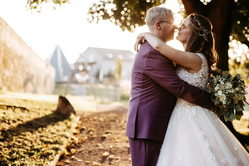 Eine Braut und ein Bräutigam umarmen sich im Freien im Sonnenlicht und lächeln sich gegenseitig an. Die Braut trägt ein weißes Kleid und hält einen Blumenstrauß in der Hand, der Bräutigam trägt einen lila Anzug. Im Hintergrund sind Bäume und ein Haus zu sehen.