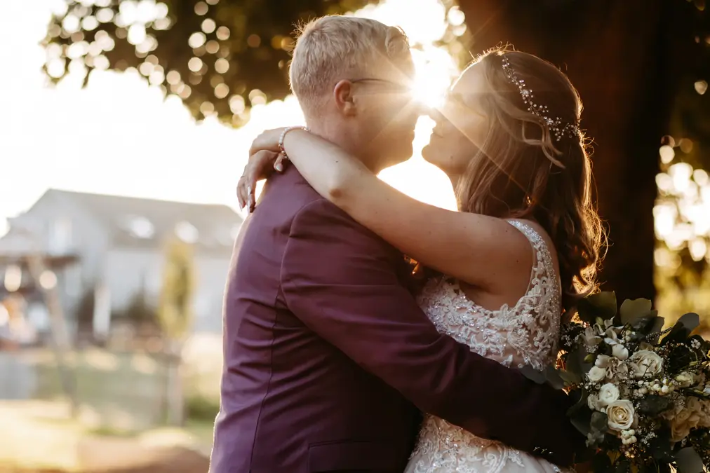 Eine Braut und ein Bräutigam umarmen sich bei Sonnenuntergang im Freien, wobei das Sonnenlicht zwischen ihren Gesichtern hindurchscheint. Die Braut hält einen Blumenstrauß und trägt ein perlenbesetztes Kleid, während der Bräutigam einen violetten Anzug trägt. Sie scheinen sich gleich küssen zu wollen.