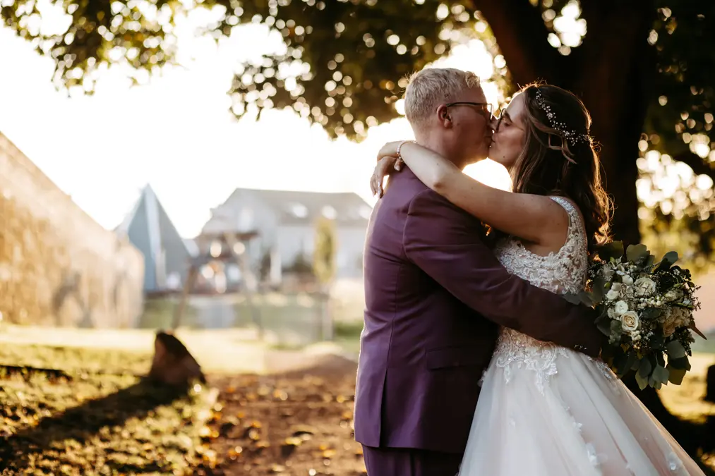Eine Braut und ein Bräutigam umarmen und küssen sich im Freien unter einem Baum, der in goldenes Sonnenlicht getaucht ist. Die Braut trägt ein weißes Kleid und hält einen Blumenstrauß; der Bräutigam trägt einen violetten Anzug. Ein Haus und Bäume erscheinen unscharf im Hintergrund.
