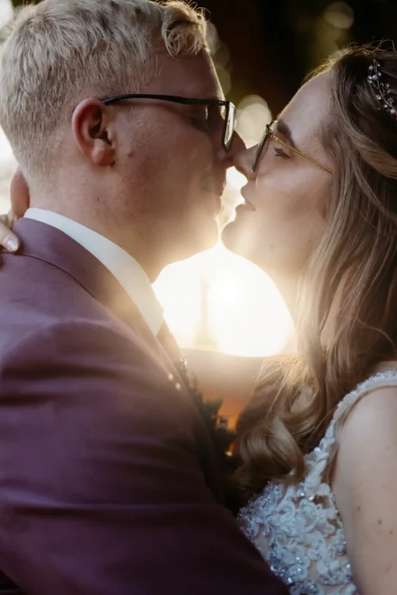 Ein Paar in Hochzeitskleidung umarmt sich eng, berührt sich an den Nasen und lächelt sanft, während das warme Sonnenlicht hinter ihnen leuchtet.