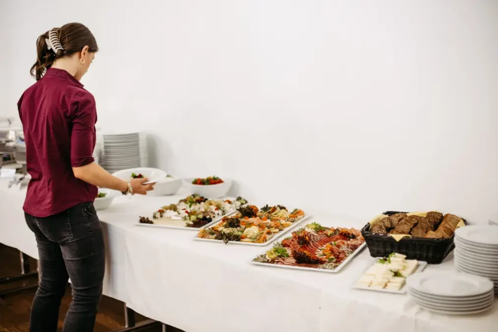 Eine Person steht an einem Büffettisch mit Tabletts mit Lebensmitteln, darunter Brot, Käse, Obst und verschiedene Gerichte. In der Nähe sind Teller und Schüsseln gestapelt. Die Person scheint das Essen auszuwählen oder zu arrangieren.