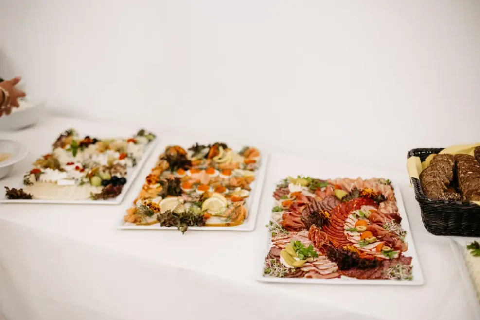 Drei Platten mit verschiedenen Vorspeisen, darunter Aufschnitt, Käse, Gemüse und Beilagen, sind auf einem weißen Tischtuch neben einem Korb mit Schwarzbrotscheiben angeordnet.