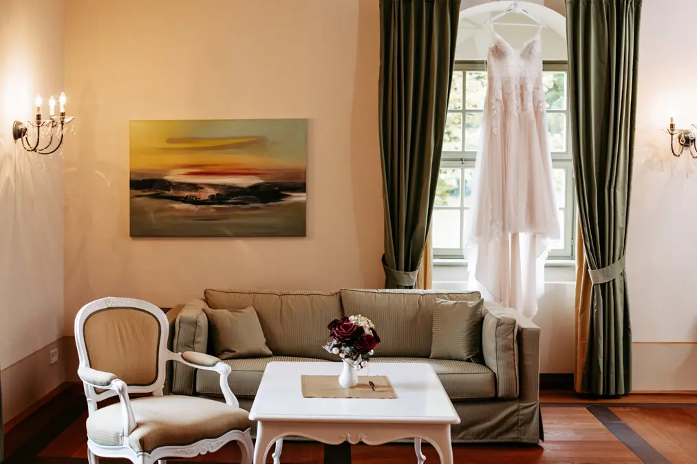 Ein weißes Hochzeitskleid hängt vor einem Fenster mit grünen Vorhängen, dahinter stehen ein beiges Sofa und ein beiger Sessel, ein weißer Couchtisch mit einem blumigen Tafelaufsatz und ein Landschaftsgemälde an der Wand.