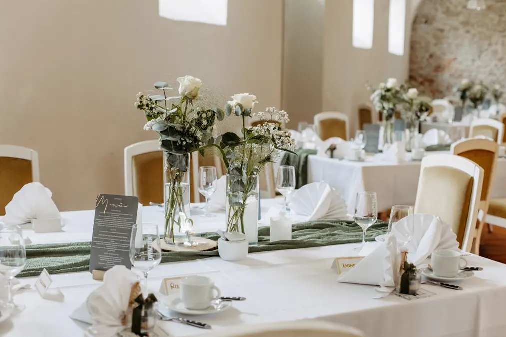 Die eleganten Banketttische sind mit weißer Bettwäsche, gefalteten Servietten, Gläsern und kleinen Blumensträußen mit weißen Blumen und Grünzeug gedeckt. Tischkarten und eine Speisekarte sind in einem sanft beleuchteten Raum mit cremefarbenen Wänden zu sehen.