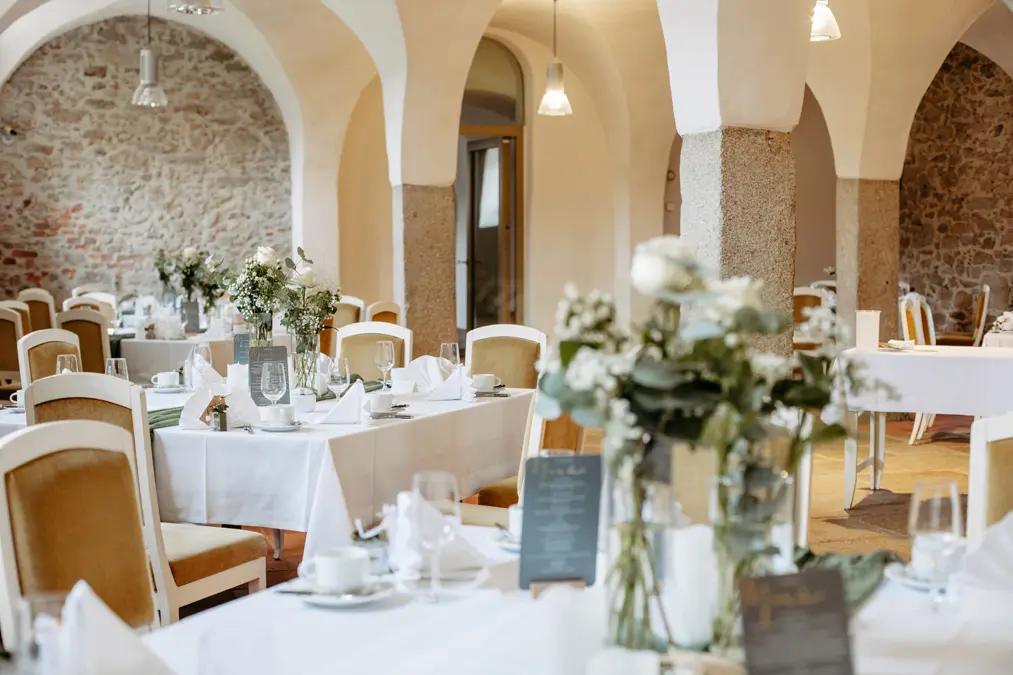 Eleganter Restaurantbereich mit gewölbten Decken, Steinwänden und weiß gedeckten Tischen, Blumenschmuck, Glaswaren und Speisekarten. Die sanfte Beleuchtung schafft eine warme, einladende Atmosphäre.