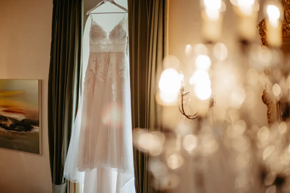 Ein weißes Spitzenhochzeitskleid hängt auf einem Kleiderbügel vor einem Fenster mit dunklen Vorhängen, das vom Sonnenlicht sanft beleuchtet wird. Ein verschwommener Kronleuchter und Wandkunst erscheinen im Vordergrund.