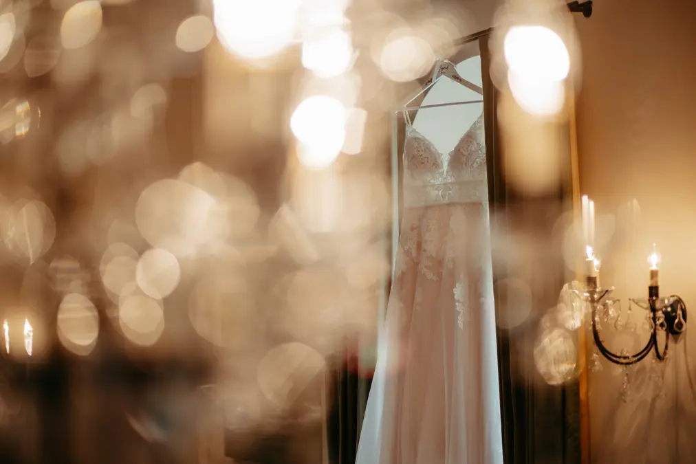 Ein weißes Spitzenhochzeitskleid hängt an einem Spiegel, der von Kerzen und verschwommenen Lichtern im Vordergrund sanft beleuchtet wird und eine verträumte, romantische Atmosphäre schafft.
