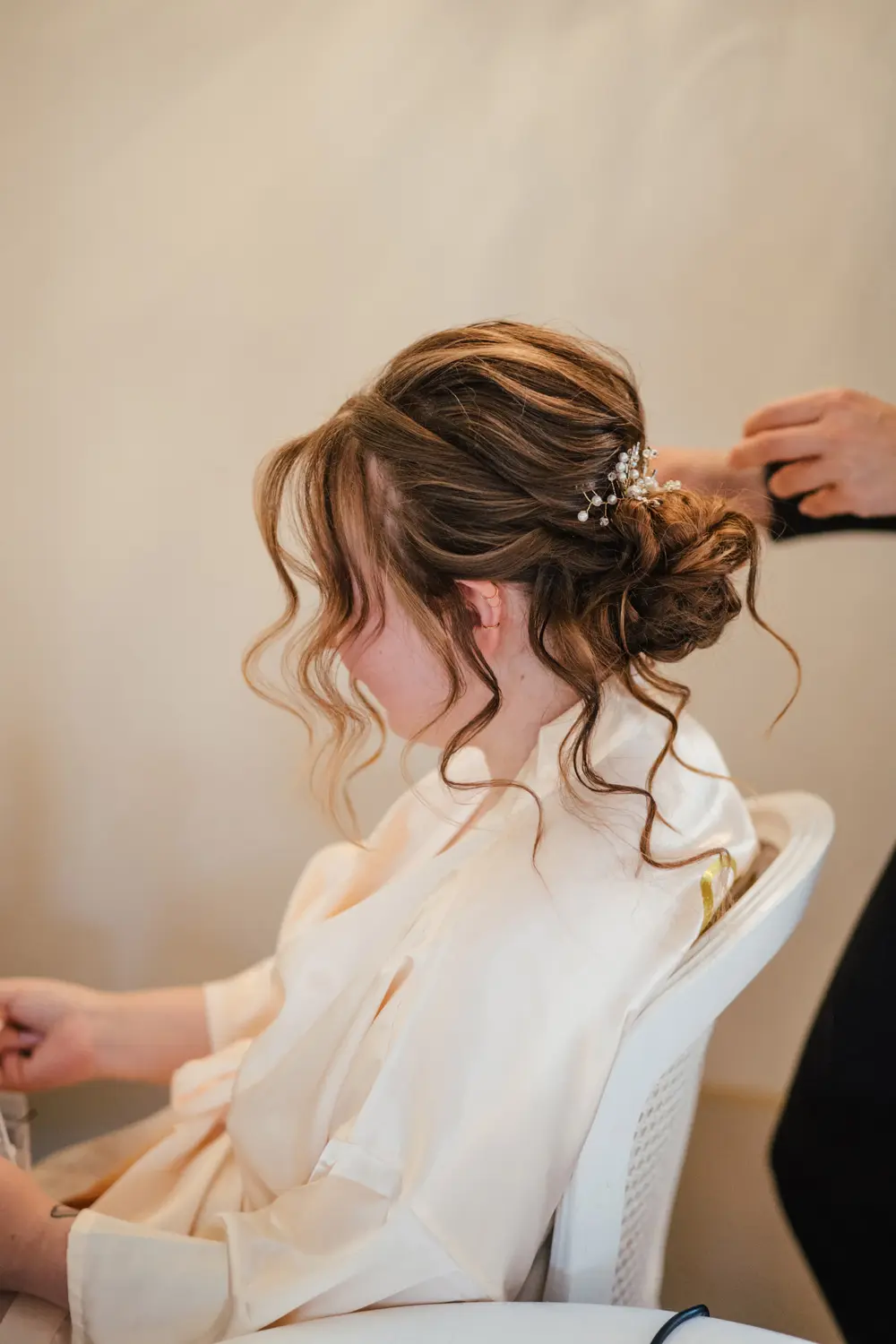 Eine Frau mit hellbraunem, gewelltem Haar, das zu einer eleganten Hochsteckfrisur mit filigranem Haarschmuck frisiert ist, sitzt in einem weißen Stuhl und trägt einen cremefarbenen Bademantel, während jemand von hinten ihre Frisur zurechtrückt.