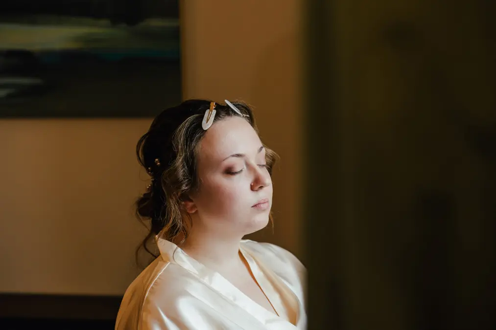 Eine Frau mit geschlossenen Augen sitzt in sanftem Licht, trägt ein weißes Gewand und Haarspangen in ihrem gewellten braunen Haar und wirkt ruhig und entspannt in ihrem Zimmer.