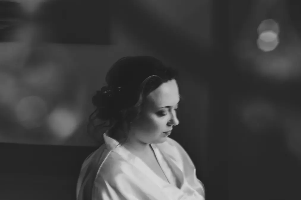 Ein Schwarz-Weiß-Foto einer Frau mit gelocktem Haar, die nach unten und leicht wegschaut und ein helles Gewand trägt, vor einem Hintergrund mit Weichzeichner.