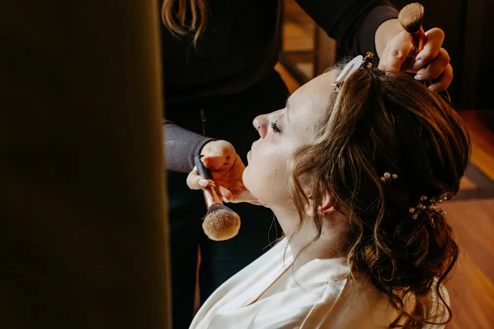 Eine Frau mit gelocktem Haar und dekorativen Haarnadeln lässt sich von einer anderen Person schminken, die mit einem Pinsel Puder auf ihr Gesicht aufträgt. Die Frau trägt einen weißen Bademantel und sitzt mit geschlossenen Augen da.