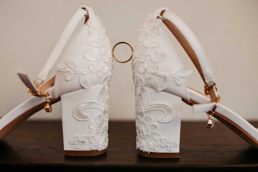 Zwei weiße, bestickte Stöckelschuhe stehen Ferse an Ferse, und zwischen ihnen balanciert ein goldener Ehering. Die Schuhe haben florale Spitzenmuster und Knöchelriemen mit goldenen Schnallen.