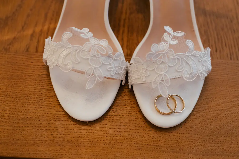 Ein Paar weißer, spitzenverzierter Brautschuhe steht auf einer hölzernen Unterlage, wobei auf dem rechten Schuh zwei goldene Eheringe ruhen.