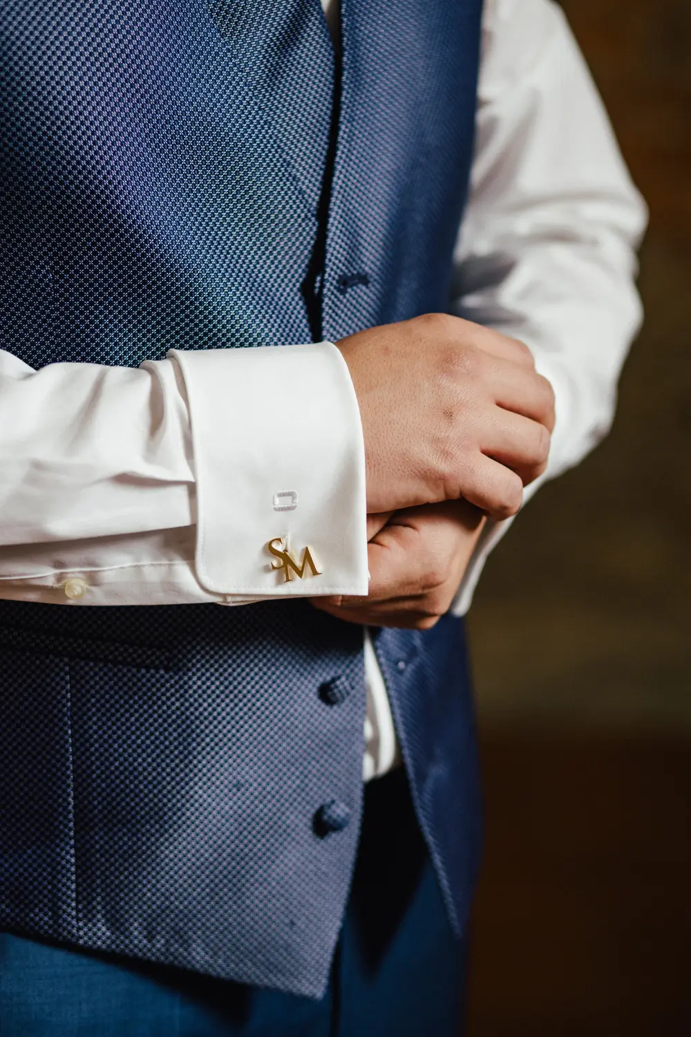 Eine Person, die eine strukturierte blaue Weste und ein weißes Hemd trägt, rückt ihren goldenen Manschettenknopf zurecht, der die Form der Buchstaben SM hat.