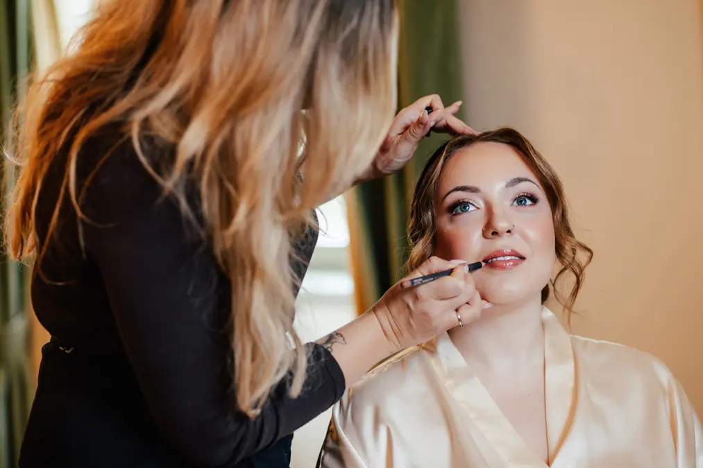 Eine Visagistin trägt Lippenstift auf eine Frau mit hellbraunem Haar auf, die ein cremefarbenes Gewand trägt und mit einem leichten Lächeln nach oben schaut. Im Hintergrund sind grüne Vorhänge und sanfte Beleuchtung zu sehen.
