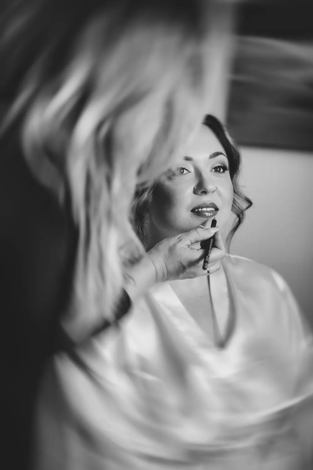 Eine Frau in einem Satinmantel lächelt, während eine andere Person ihren Lippenstift aufträgt. Das Bild ist schwarz-weiß, mit einem weichen, unscharfen Effekt, der eine verträumte, intime Atmosphäre schafft.