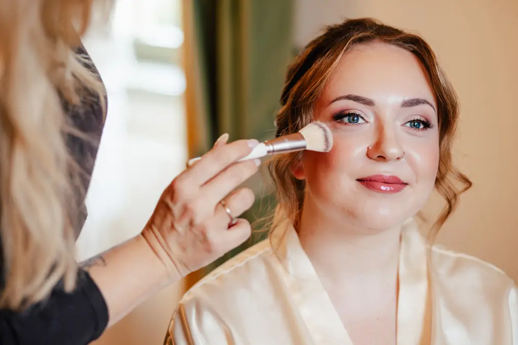 Eine Frau mit hellen Augen und gestyltem Haar lächelt sanft, während eine andere Person mit einem Pinsel Make-up auf ihre Wangen aufträgt, um sie auf einen besonderen Anlass vorzubereiten.