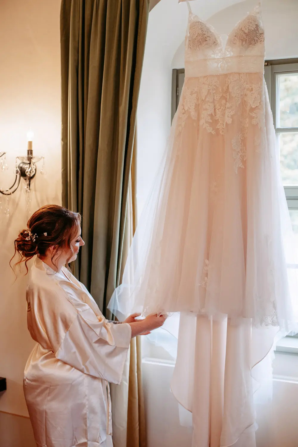 Eine Frau in einem weißen Gewand steht an einem Fenster, lächelt und berührt sanft ein langes, weißes Hochzeitskleid aus Spitzenstoff, das aufgehängt ist.