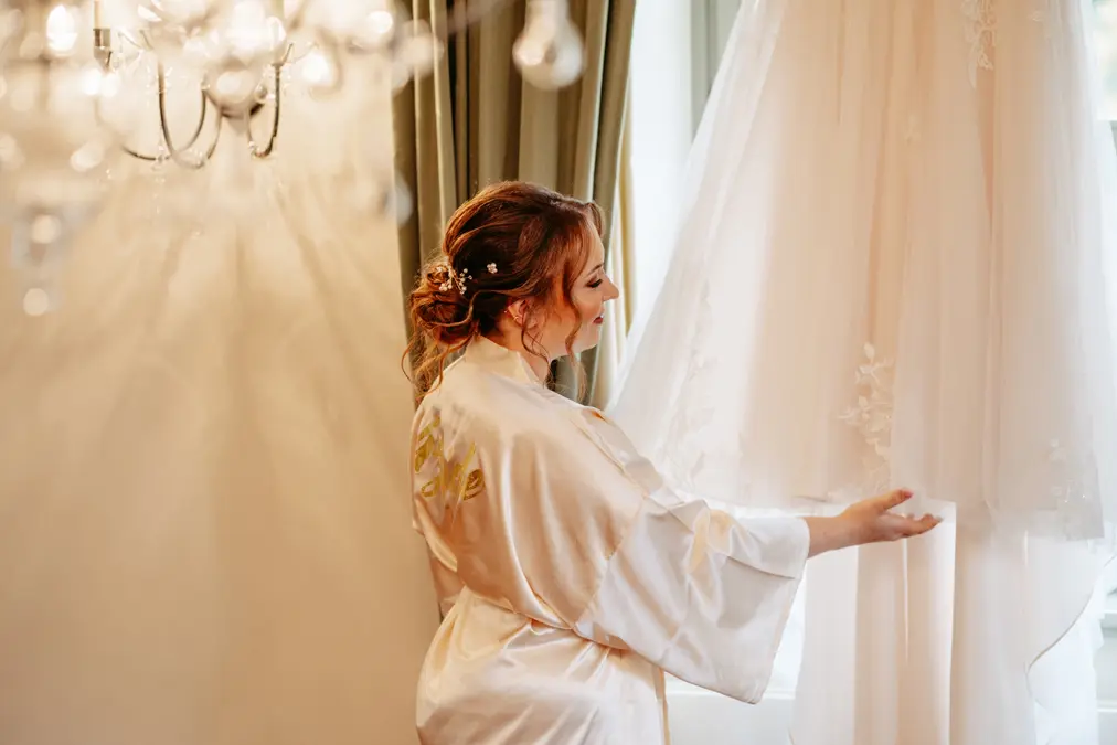 Eine Frau in einem Satinmantel steht am Fenster und hält lächelnd den Saum eines weißen Hochzeitskleides fest, das vor ihr hängt. Elegante Beleuchtung und Vorhänge verleihen dem Raum eine weiche, warme Atmosphäre.