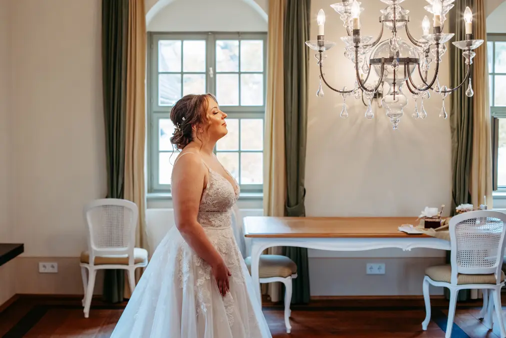 Eine Braut in einem weiĂźen Spitzenhochzeitskleid steht in einem sanft beleuchteten Raum und blickt auf ein helles Fenster. Der Raum ist mit eleganten StĂĽhlen, einem Holztisch und einem KristalllĂĽster ĂĽber dem Kopf ausgestattet.