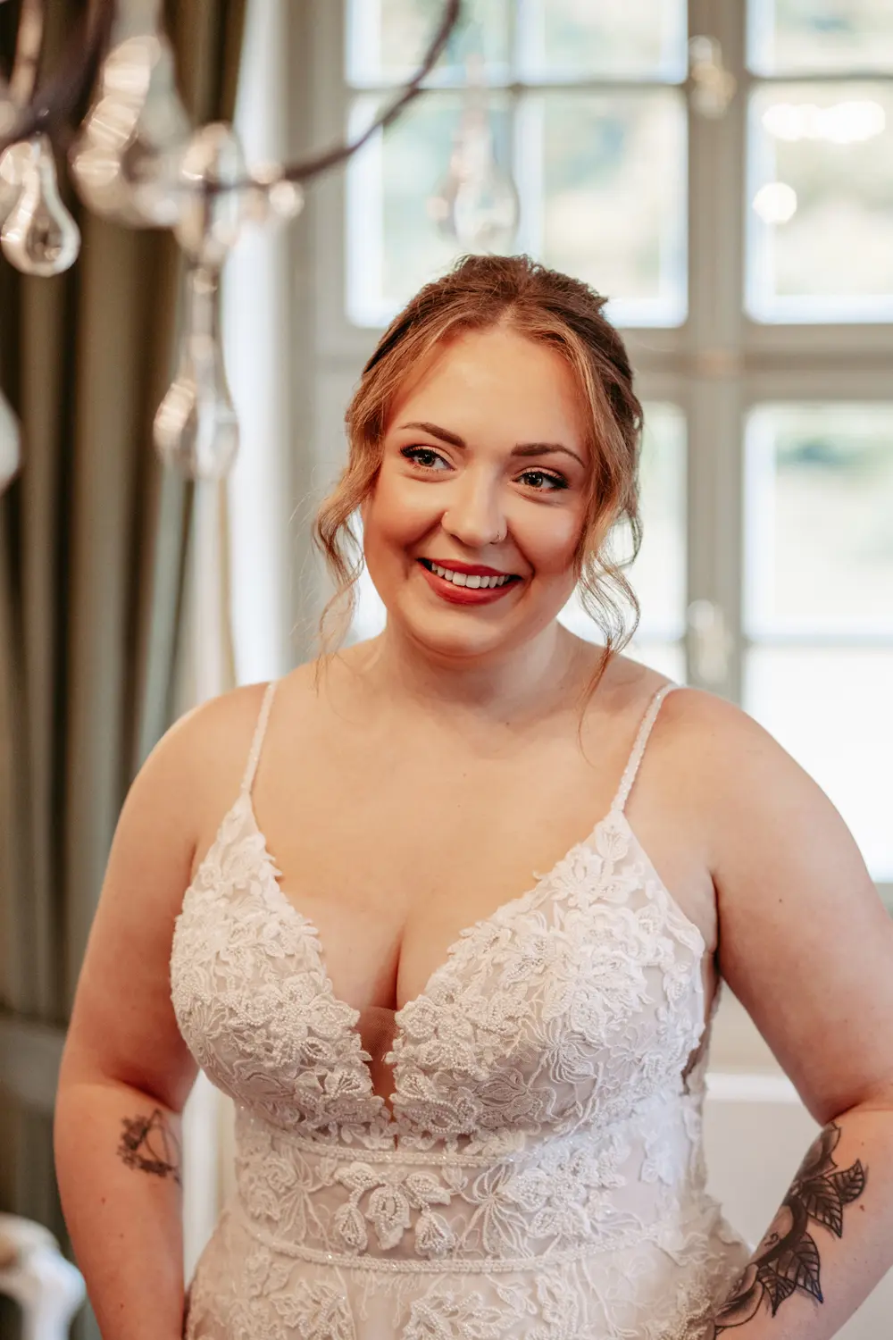 Eine lächelnde Frau mit hellbraunem Haar in einem weißen Spitzenhochzeitskleid steht in einem Innenraum in der Nähe eines Fensters, wobei weiches natürliches Licht ihr Gesicht und ihre Schultern beleuchtet.