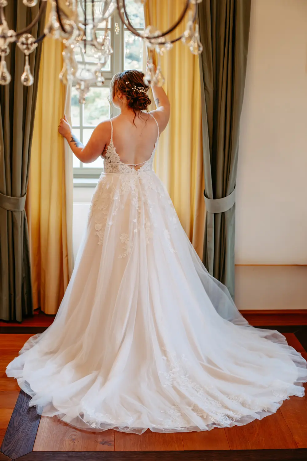 Eine Braut in einem weißen Hochzeitskleid aus Spitze steht mit dem Rücken zur Kamera und blickt aus einem Fenster, das von gelben und grünen Vorhängen eingerahmt wird. Ihr Haar ist hochgesteckt und sie hält einen Vorhang vorsichtig zurück.