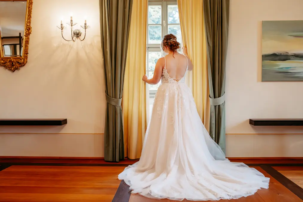 Eine Braut in einem weißen Hochzeitskleid steht auf einem Holzboden vor einem Fenster mit grünen und gelben Vorhängen und berührt sanft den Stoff. Licht strömt herein und beleuchtet den eleganten Raum mit einem Wandleuchter, einem Spiegel und einem Gemälde.