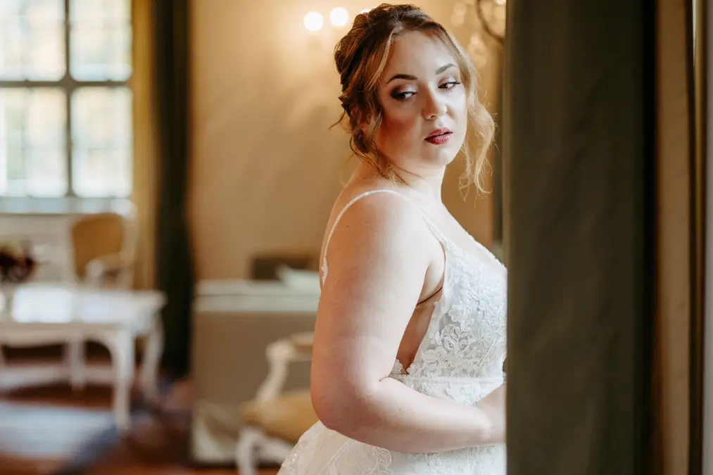 Eine Braut in einem weißen Spitzenhochzeitskleid steht in einem Raum und schaut über ihre Schulter in Richtung Kamera. Der sanft beleuchtete Raum ist mit eleganten Möbeln und großen Fenstern im Hintergrund ausgestattet.