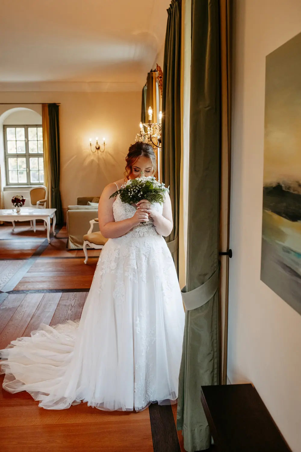 Eine Braut in einem weißen Spitzenkleid steht drinnen an einem Fenster und hält sich einen Blumenstrauß vors Gesicht. Der elegante Raum ist mit Holzböden, grünen Vorhängen und klassischen Möbeln ausgestattet.