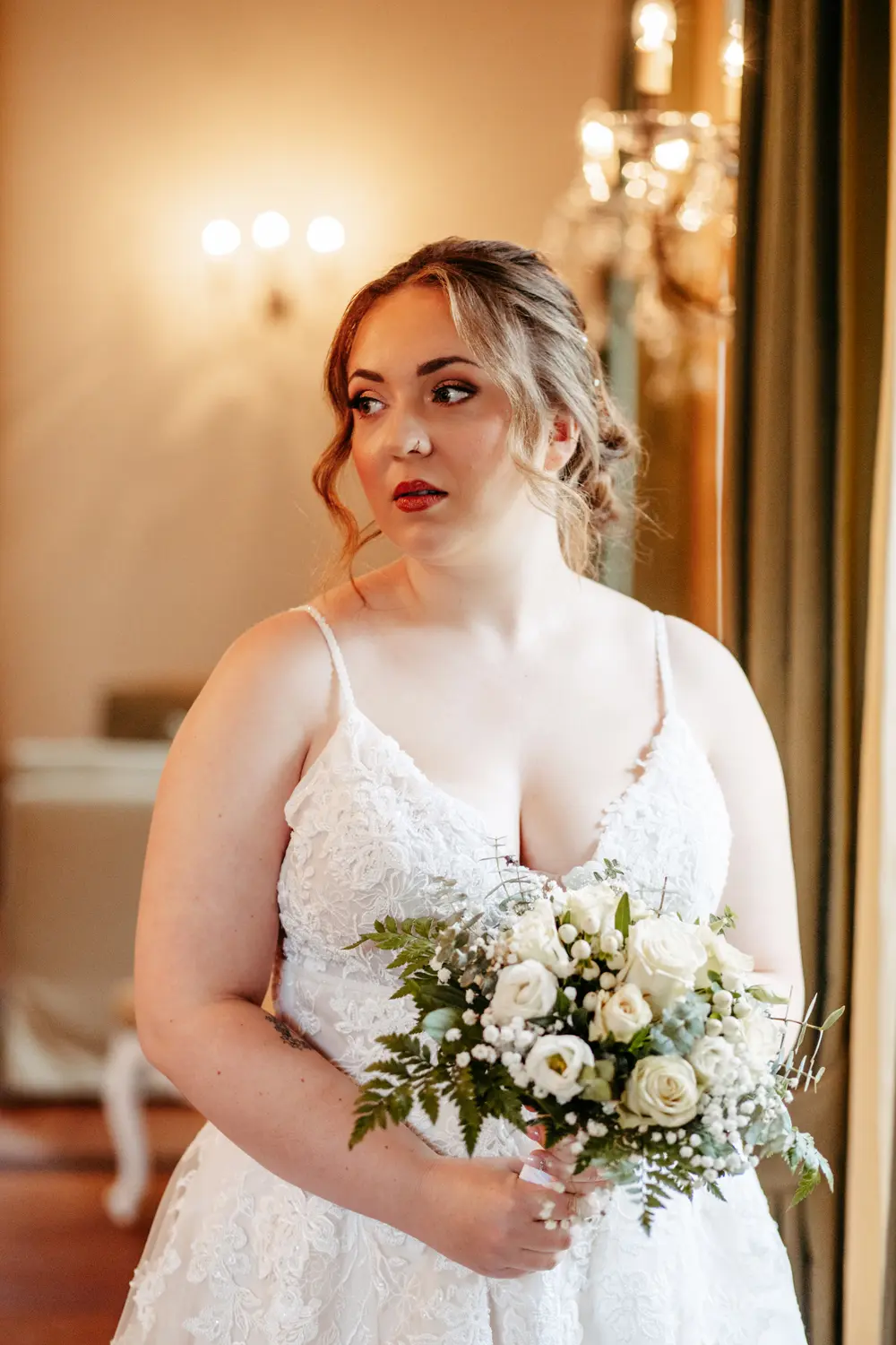 Eine Braut in einem weißen Spitzenhochzeitskleid hält einen Strauß weißer Rosen und Grünzeug in der Hand. Sie steht in einem Innenraum in der Nähe eines Fensters mit eleganter Beleuchtung und Dekoration im Hintergrund.