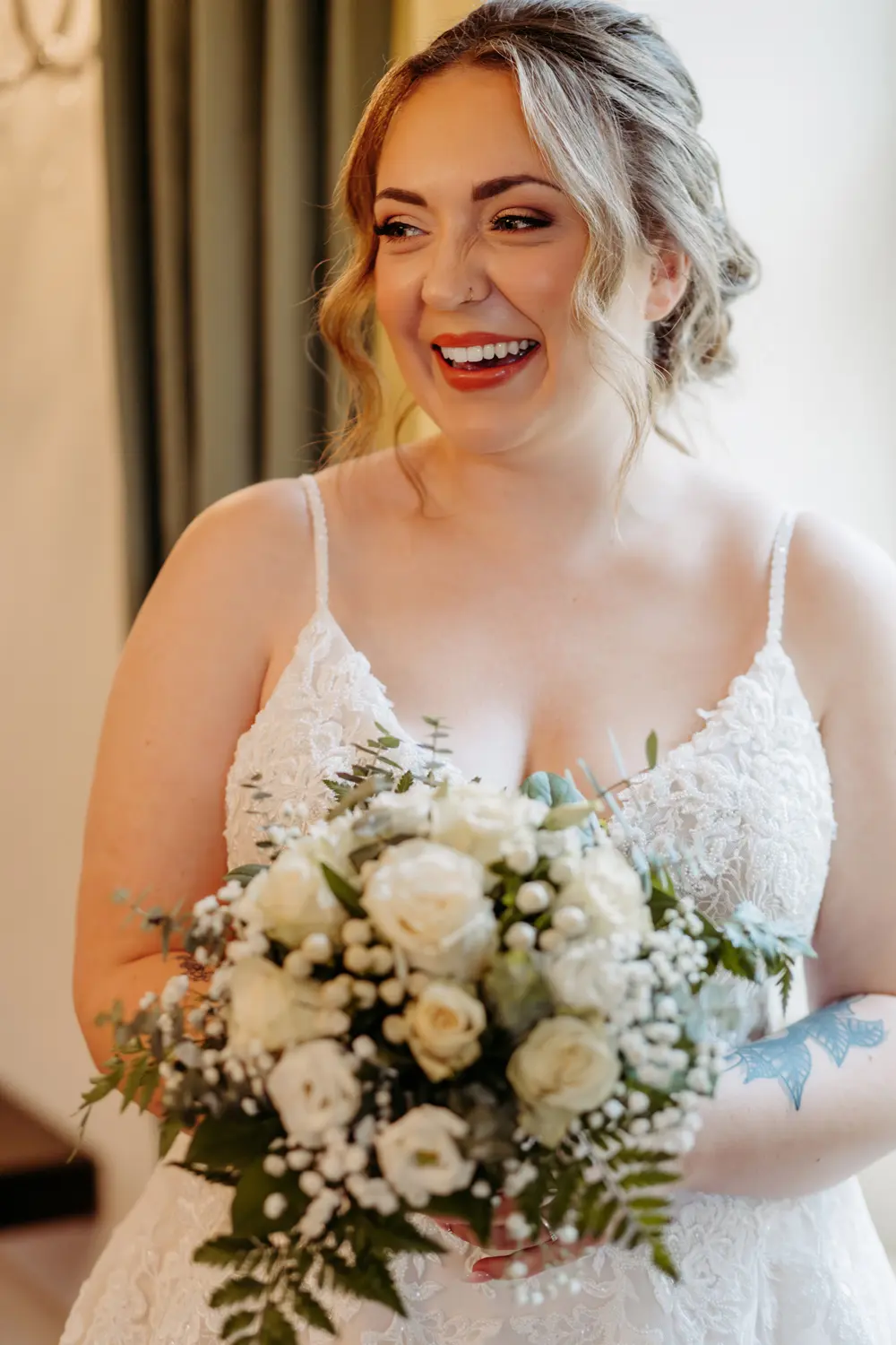 Eine lächelnde Braut in einem weißen Spitzenkleid hält einen Strauß aus weißen Rosen und Grünzeug. Sie hat ihr gewelltes Haar hochgesteckt und steht in einem Innenraum mit grünen Vorhängen im Hintergrund.