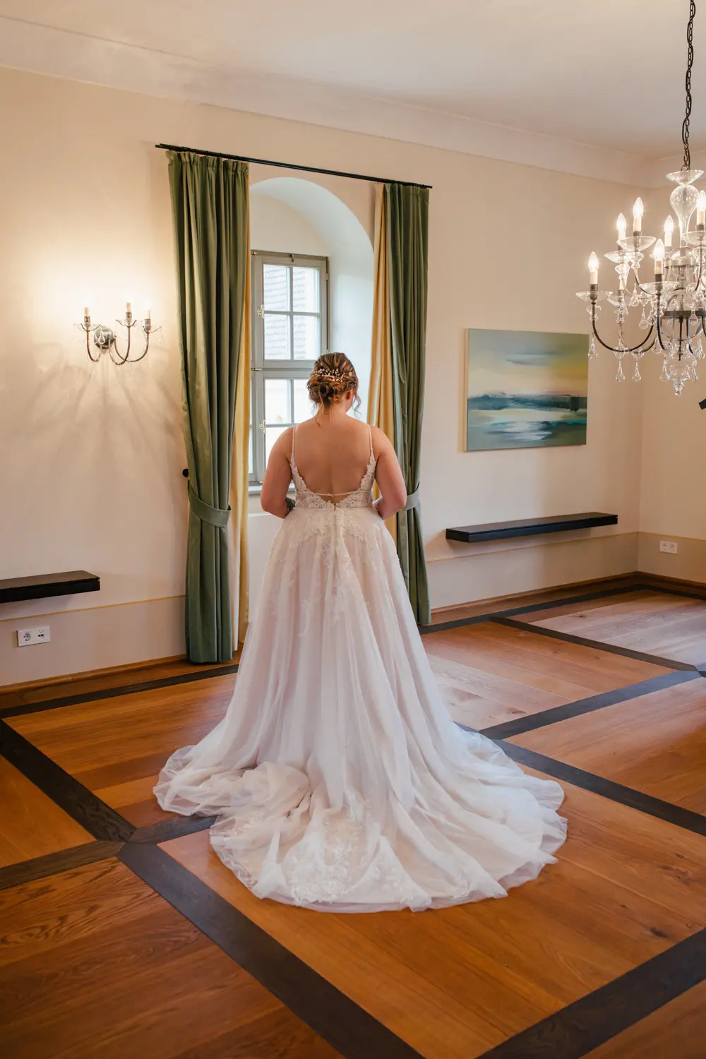 Eine Braut in einem weißen Hochzeitskleid steht mit dem Rücken zur Kamera in einem hellen Raum mit Holzfußboden, grünen Vorhängen, Wandleuchten und einem Kronleuchter.