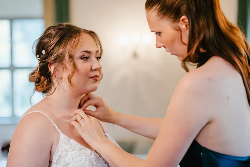 Eine Frau in einem weißen Spitzenkleid steht still, während ihr eine andere Frau dabei hilft, eine Halskette um ihren Hals zu legen. Beide wirken in einem schwach beleuchteten Raum konzentriert.