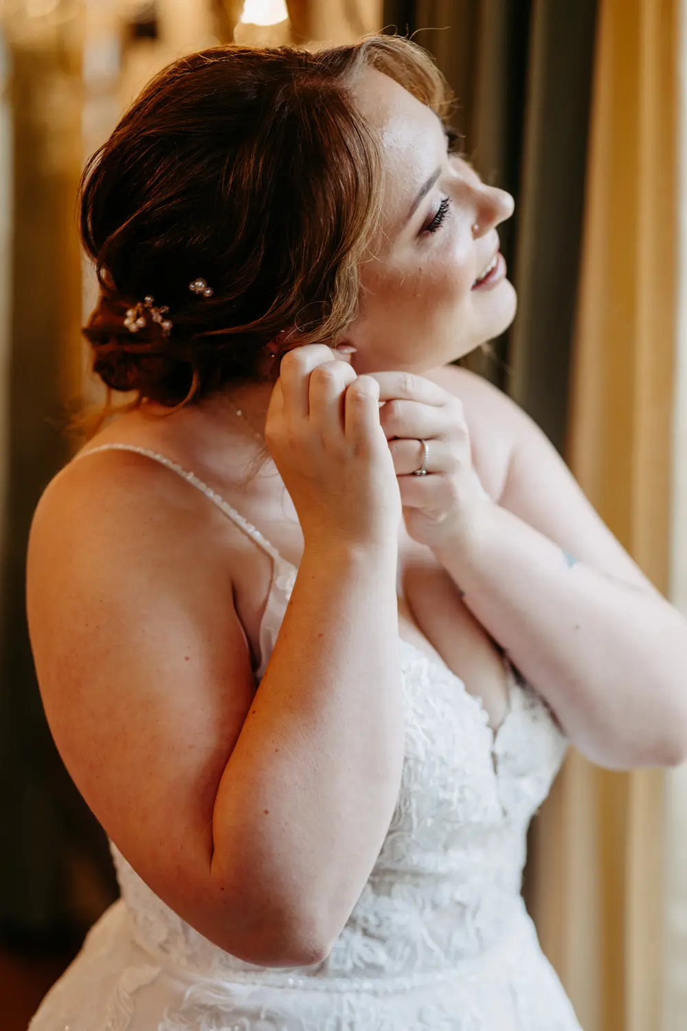Eine Braut mit kastanienbraunem Haar in einer Hochsteckfrisur rückt ihren Ohrring zurecht, während sie lächelt und zur Seite schaut. Sie trägt ein weißes Spitzenhochzeitskleid mit dünnen Trägern. Weiches, warmes Licht erhellt ihr Gesicht.