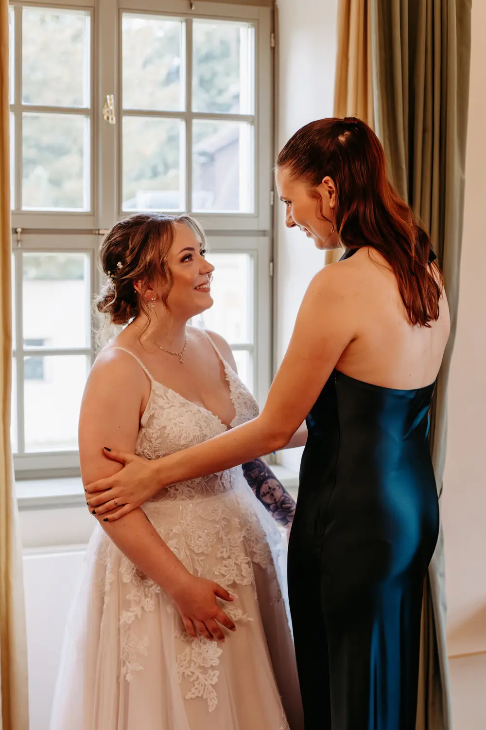 Zwei Frauen stehen an einem Fenster in einem Innenraum, die eine in einem weißen Spitzenhochzeitskleid, die andere in einem dunklen, rückenfreien Kleid. Sie schauen sich an und lächeln sich warm zu, teilen einen liebevollen, unterstützenden Moment.