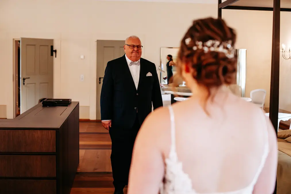 Ein Mann in Anzug und Fliege lächelt, während er eine Frau in einem weißen Kleid ansieht, die mit dem Rücken zur Kamera steht, wahrscheinlich während einer Hochzeit in einem warm beleuchteten Raum.