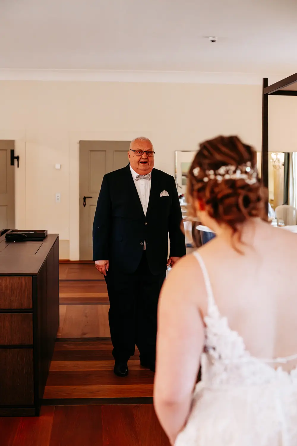 Ein Mann im Anzug lächelt freundlich, als er eine Frau in einem weißen Hochzeitskleid mit geblümtem Haarschmuck sieht, die mit dem Rücken zur Kamera in einem Raum mit Holzboden und neutralen Wänden steht.
