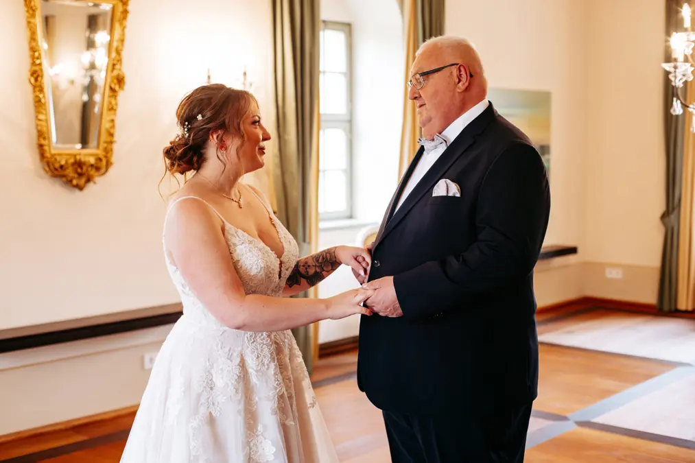 In einem hellen, eleganten Raum mit Holzböden, großen Fenstern und goldgerahmten Spiegeln hält eine Braut in einem weißen Hochzeitskleid die Hände und lächelt einem älteren Mann im Anzug zu.