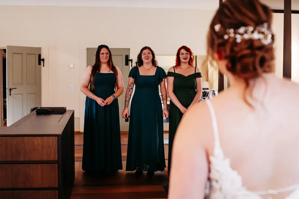 Drei Frauen in grünen Kleidern lächeln eine Braut in einem weißen Kleid an, die mit dem Rücken zur Kamera steht. Sie stehen in einem gut beleuchteten Raum mit Holzboden und neutralen Wänden und verbringen einen fröhlichen Moment vor der Zeremonie.